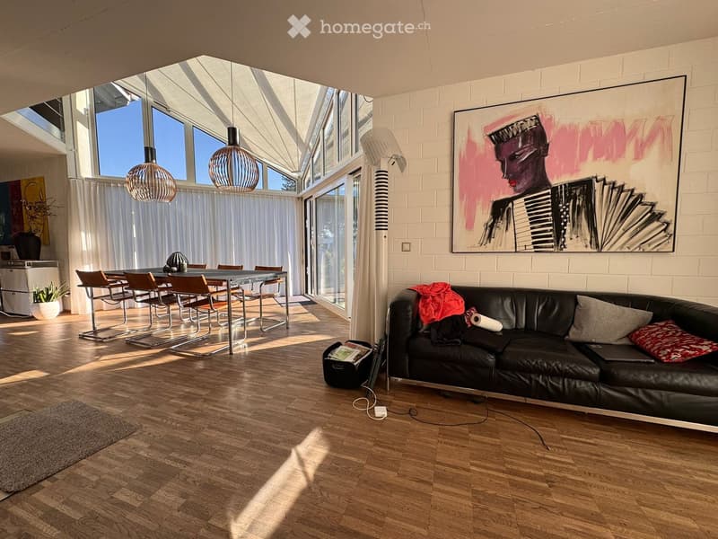 Modern living room with wooden floor, large windows, and a dining area. Features a black leather couch, dining table, and chairs. A large painting adorns the wall, and a floor lamp is placed beside the couch. A rug and a potted plant add to the decor.