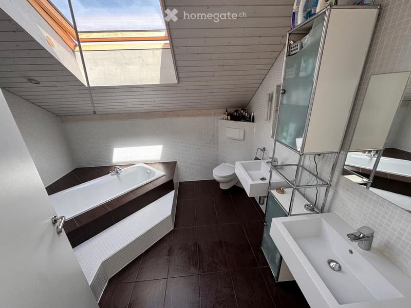 Modern bathroom with a bathtub, toilet, two sinks, and a large mirror. The room features wooden slatted ceilings and dark tiled floors.