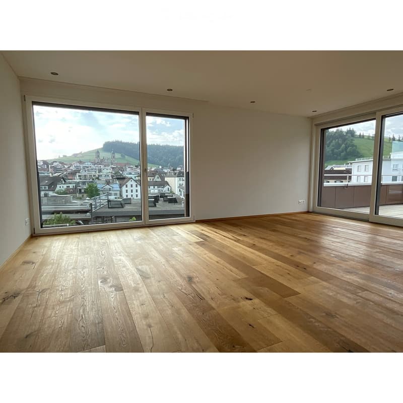 Empty room with wooden flooring and large windows offering a scenic view of a town and surrounding landscape.