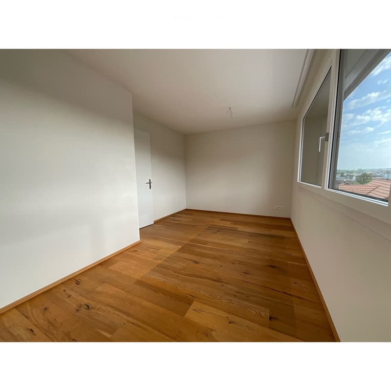Empty room with wooden flooring, white walls, and large windows offering a view of the cityscape.