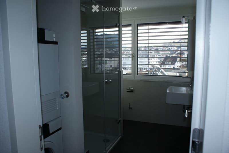 Modern bathroom with a glass-enclosed shower, a sink, and a toilet. Large windows with blinds offer a view of the city.