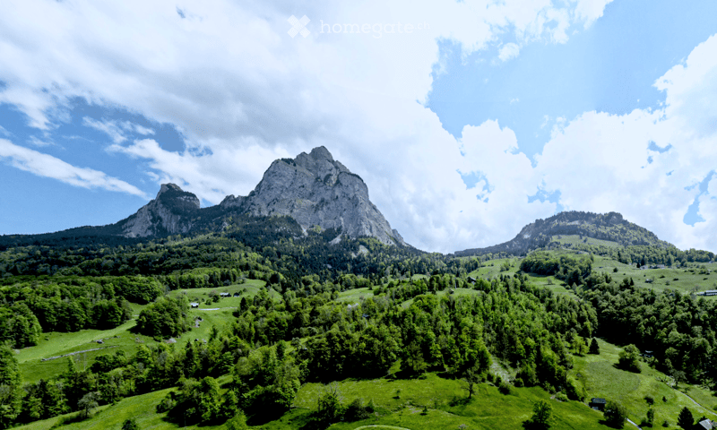 A panoramic view of a lush green valley surrounded by towering mountains under a partly cloudy sky. The landscape features dense forests, rolling hills, and scattered small houses or cabins nestled among the trees.