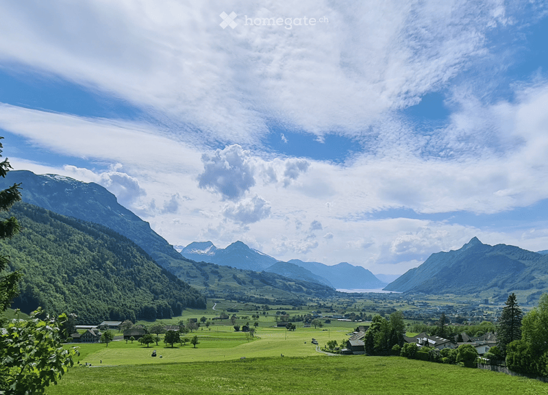 A panoramic view of a lush green valley surrounded by snow-capped mountains, with scattered houses and trees, under a partly cloudy sky.
