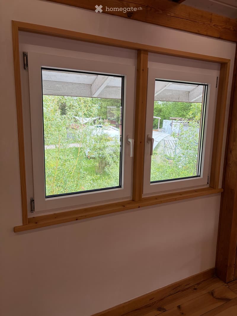 Empty room with light pink walls, wooden floor, and two windows with wooden frames and sills, offering a view of greenery and a covered outdoor area.