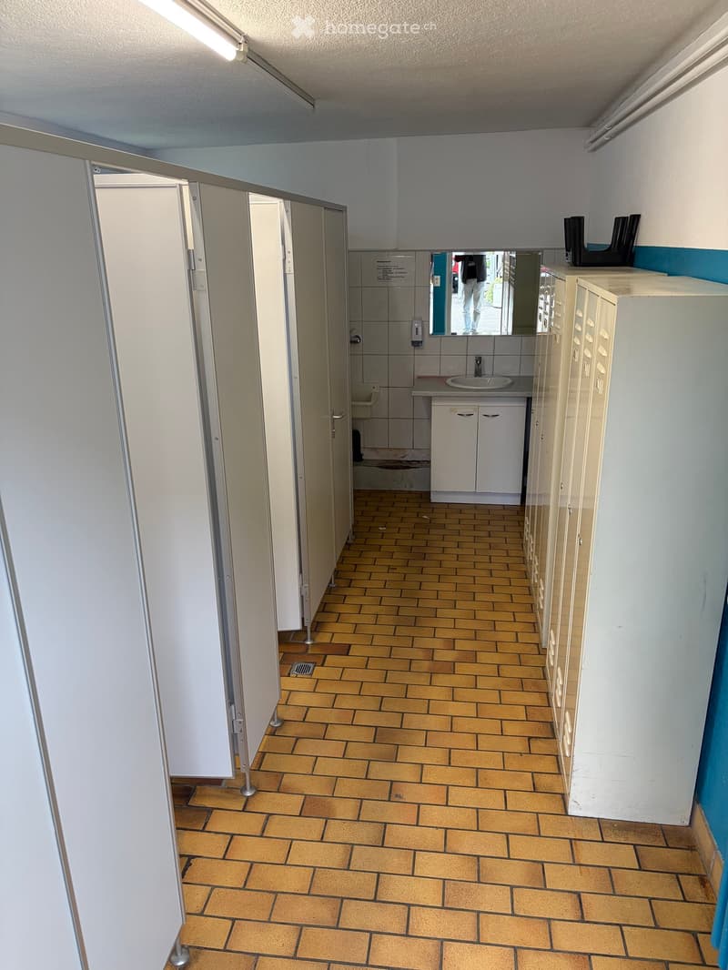 Empty hallway with tiled floor, white walls, and open doors leading to individual stalls. The area includes a sink and mirror in the background.