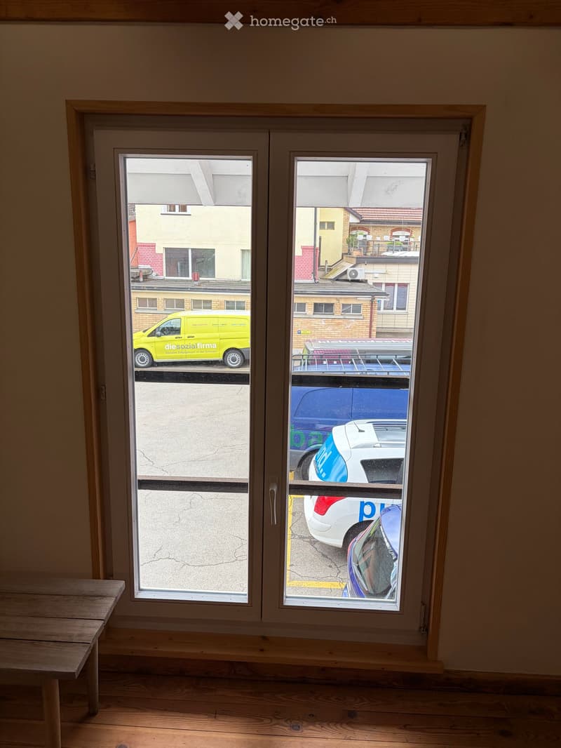 Empty room with wooden flooring and large windows showing a view of the street and parked cars.