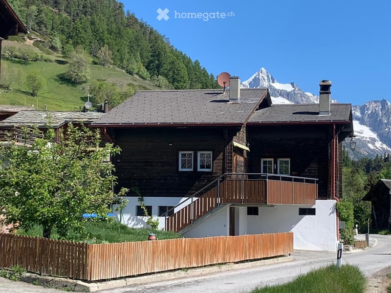 A two-story wooden house with a balcony, surrounded by a wooden fence and set against a backdrop of green hills and snow-capped mountains.