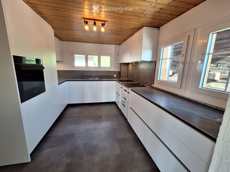 Modern kitchen with white cabinets, dark countertops, and built-in appliances including an oven and stove. Features a wooden ceiling with integrated lighting and large windows allowing natural light.