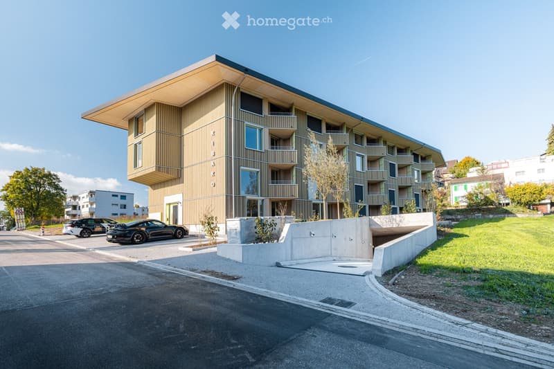 Modern multi-story building with a sleek, contemporary design featuring large windows and balconies. The structure has a combination of wood and metal finishes, with a prominent entrance and a raised ground floor area.