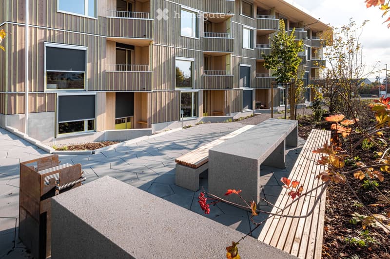 Modern multi-story building with wooden and concrete exterior, featuring balconies and large windows. The foreground shows a paved area with benches and small garden beds.