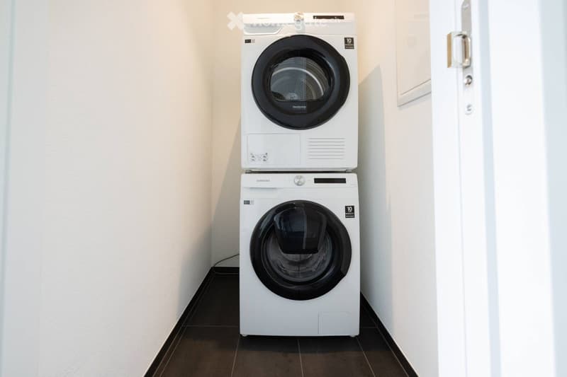 A compact room with a stacked washing machine and dryer unit, white walls, and dark tiled flooring.
