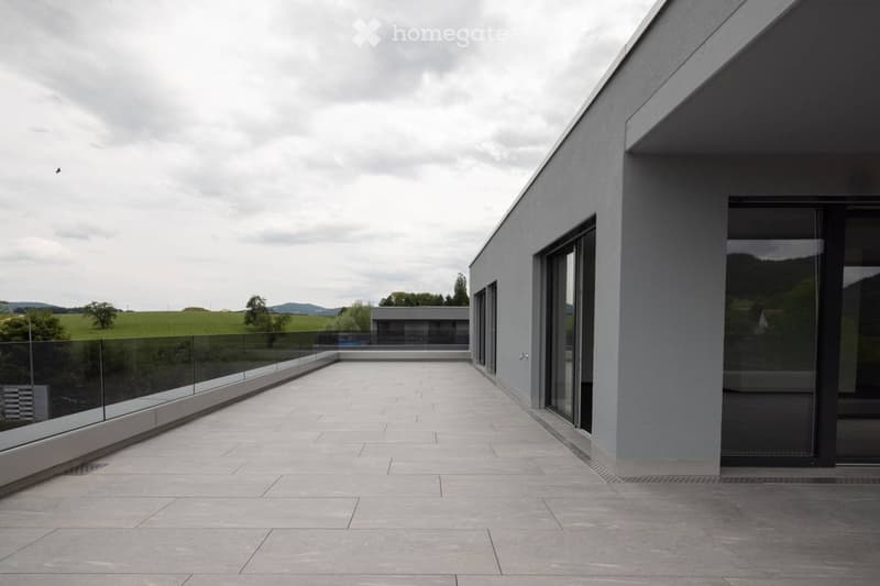 Modern balcony with large glass railings, tiled floor, and expansive view of green fields and hills.