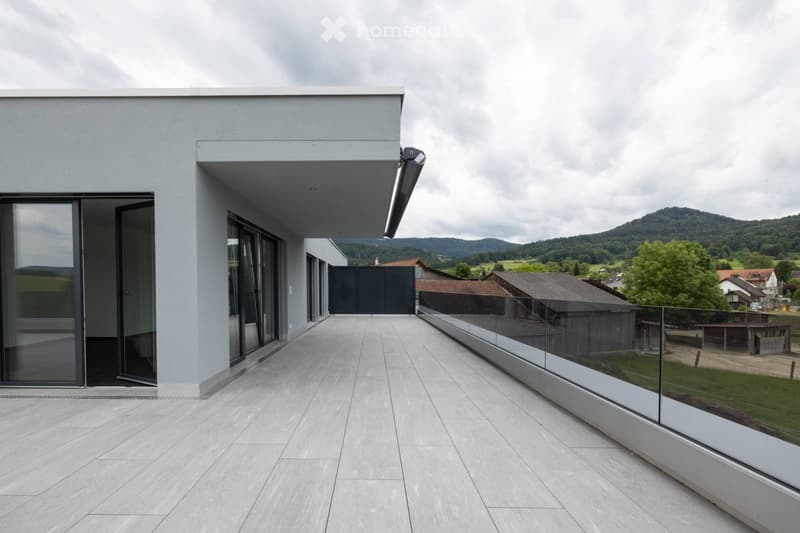 Modern balcony with large glass sliding doors, sleek grey tiles, and a glass railing overlooking a scenic countryside view.