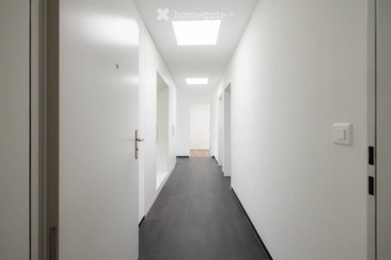 Modern, minimalist hallway with white walls, grey tiled floor, and recessed ceiling lights. Features several closed white doors with silver handles on both sides.