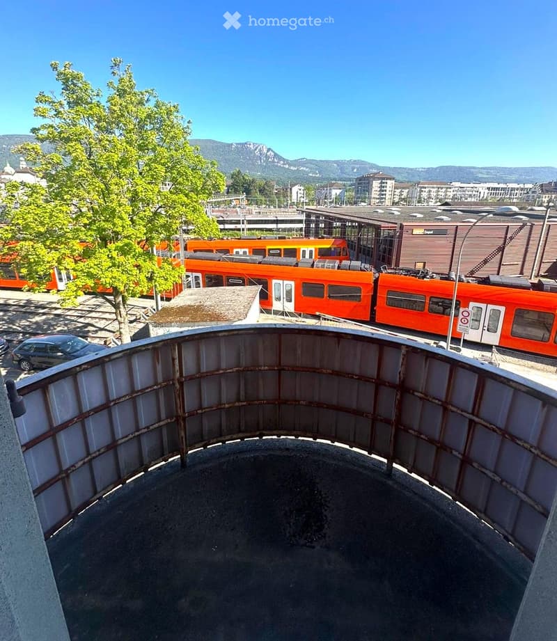 View from a rooftop terrace showing a train passing by, with a tree, parked cars, and distant mountains in the background.