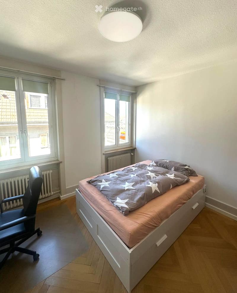 A small, well-lit bedroom with a single bed, a chair, and large windows offering a view of the street.