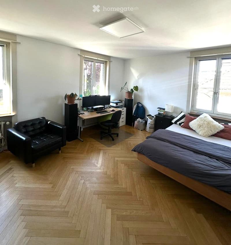 Bright bedroom with wooden flooring, large windows, a double bed with pillows and blankets, a black leather sofa, a desk with a computer, and a chair.