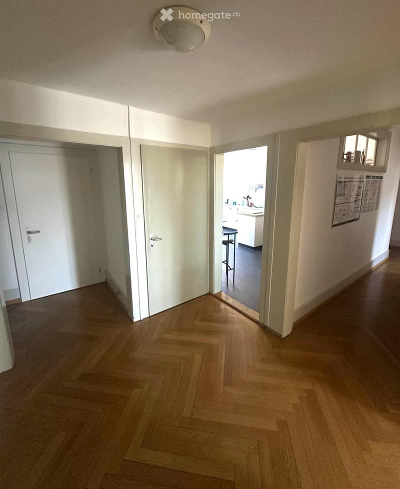A clean, well-lit hallway with wooden flooring and white walls. Three doors are visible, with one leading to a kitchen area that includes a stool and cabinets.