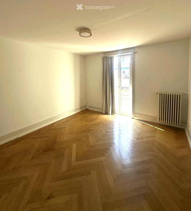 Empty room with wooden herringbone flooring, white walls, a ceiling light fixture, a radiator, and a large window with curtains leading to a balcony.