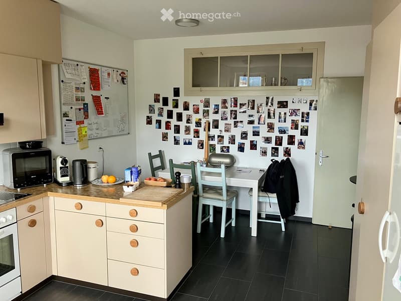A modern kitchen with light-colored cabinets, a black tiled floor, and a white refrigerator adorned with photos. The kitchen features a microwave, coffee maker, and a small dining area with a table and chairs.