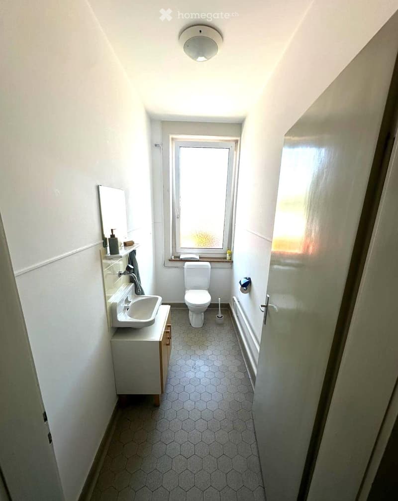 Small bathroom with hexagonal tiled floor, white walls, a sink with a mirror, a toilet, and a bathtub. A window provides natural light.