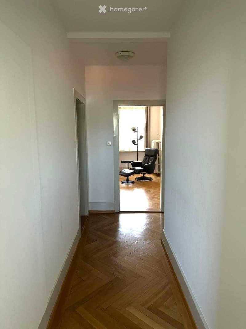 A narrow hallway with wooden flooring and white walls, leading to an adjacent room with a black leather chair and a small table.