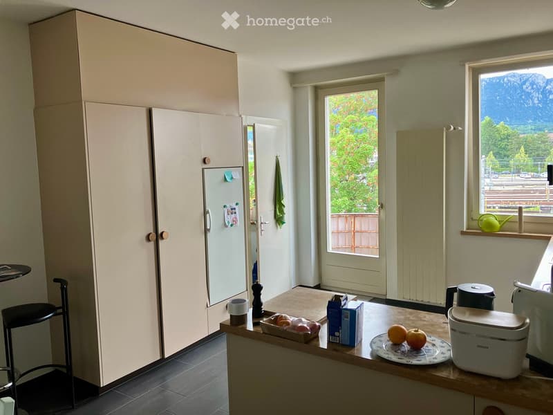 A modern kitchen with light-colored cabinets, a wooden countertop, and various appliances including a refrigerator, oven, and dishwasher. The countertop holds a cup, a bottle, a plate with fruits, and a small basket with eggs. A window offers a scenic mou