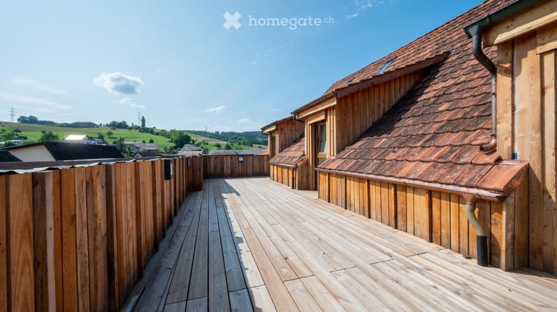 Wooden balcony with wooden decking, wooden fence, and view of a rural landscape with green fields and scattered houses.