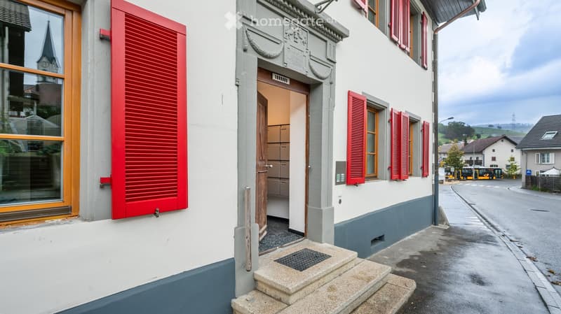 A two-story building with white walls, red shutters, and a wooden door. The entrance features a small staircase leading to the door, and the building is situated on a street with a view of a church spire in the background.