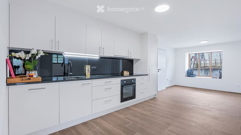 Modern kitchen with white cabinets, dark countertops, and a sink with a faucet. There is an oven, a flower arrangement, and a cutting board on the counter. The floor is wooden, and there is a window with a view of the outside.