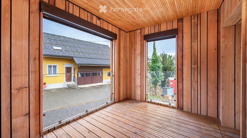 Wooden balcony with large glass doors opening to a view of a yellow house and a tree-lined street.