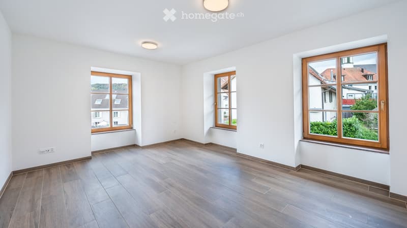 Empty room with wooden flooring, white walls, and three large windows with wooden frames, offering views of neighboring buildings and greenery.