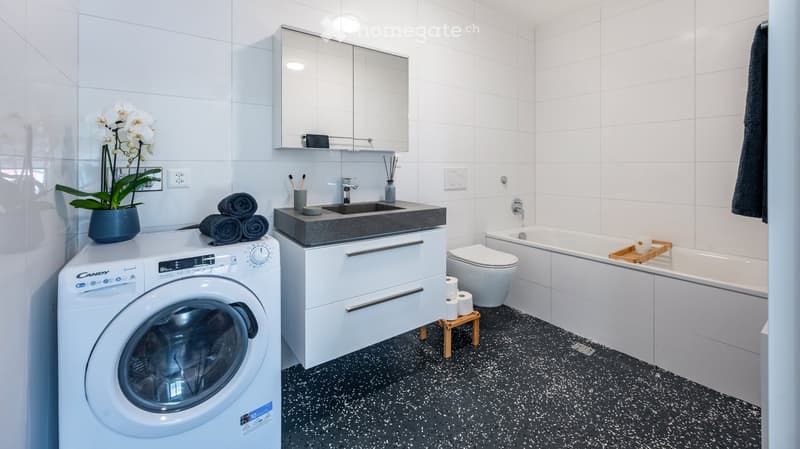 Modern bathroom with a washing machine, sink, toilet, bathtub, and a mirror cabinet. Features a black speckled floor and white tiled walls.