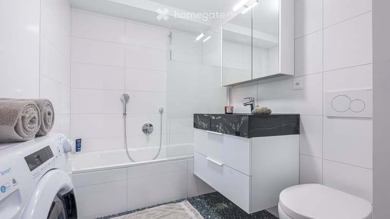 Modern bathroom with white tiles, a bathtub, a sink with a black countertop, a mirror, a toilet, and a washing machine. The space is bright with built-in lighting and features a sleek, contemporary design.