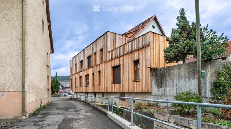 Modern wooden building with a flat roof and multiple windows, situated on a raised ground floor with a metal railing and steps leading up to the entrance.
