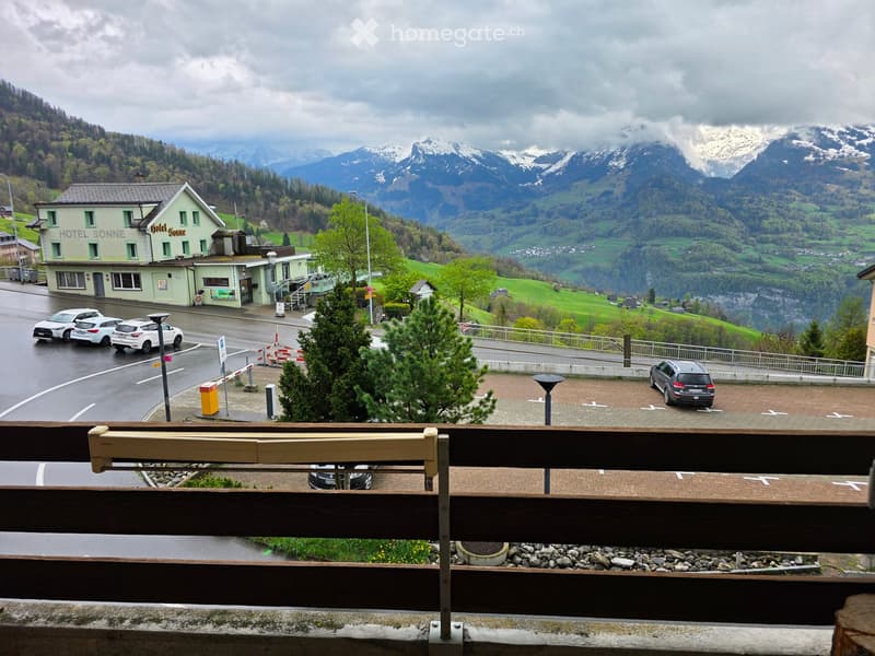View from a balcony overlooking a parking area, a hotel named "Hotel Sonne," and a scenic mountain landscape with snow-capped peaks and green valleys.