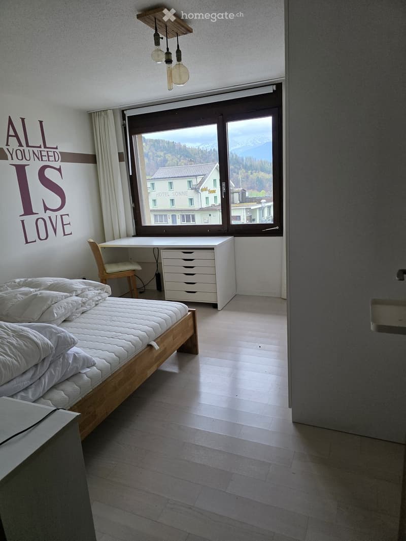 Modern bedroom with a wooden bed, desk, chair, and cabinet, featuring large windows with a scenic mountain view.