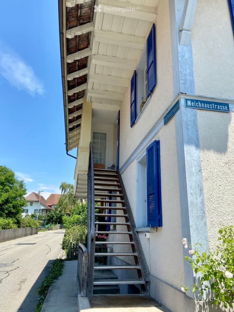 Raised ground floor house with wooden stairs, blue shutters, white exterior wall