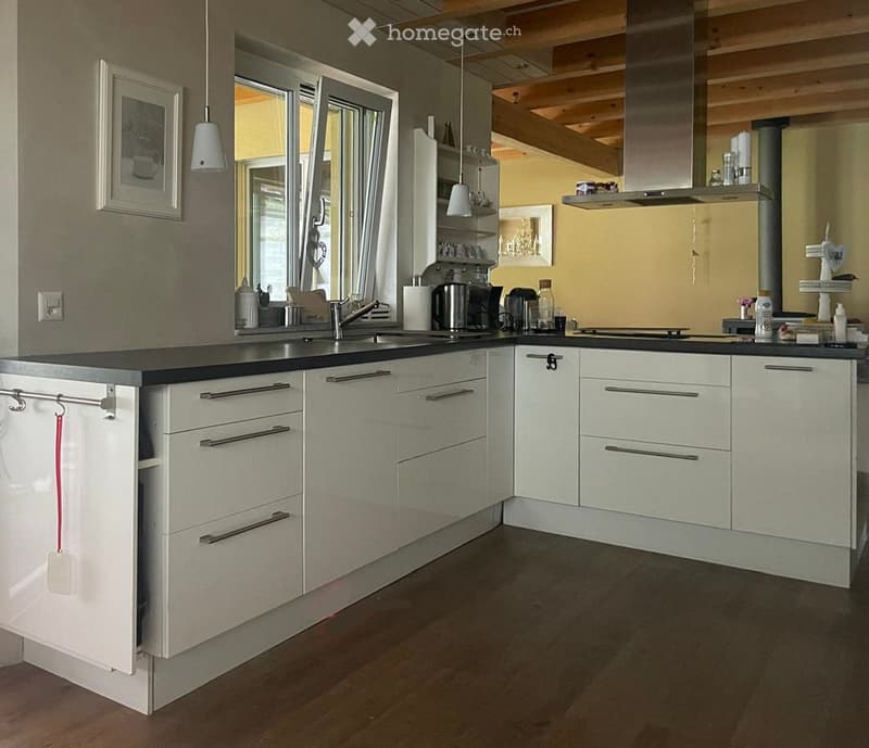 Modern kitchen with white cabinets, dark countertops, pendant lights, and various appliances including a coffee maker, microwave, and toaster. The kitchen features a central island with a sink and faucet, and there are framed pictures on the walls.