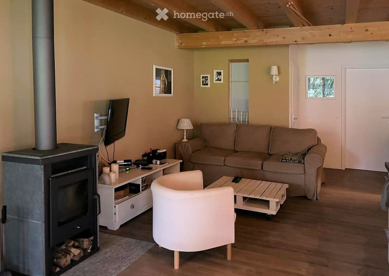 Cozy living room with exposed wooden beams, a fireplace, a beige sofa, a white armchair, a wooden coffee table, a television, and a small table lamp.