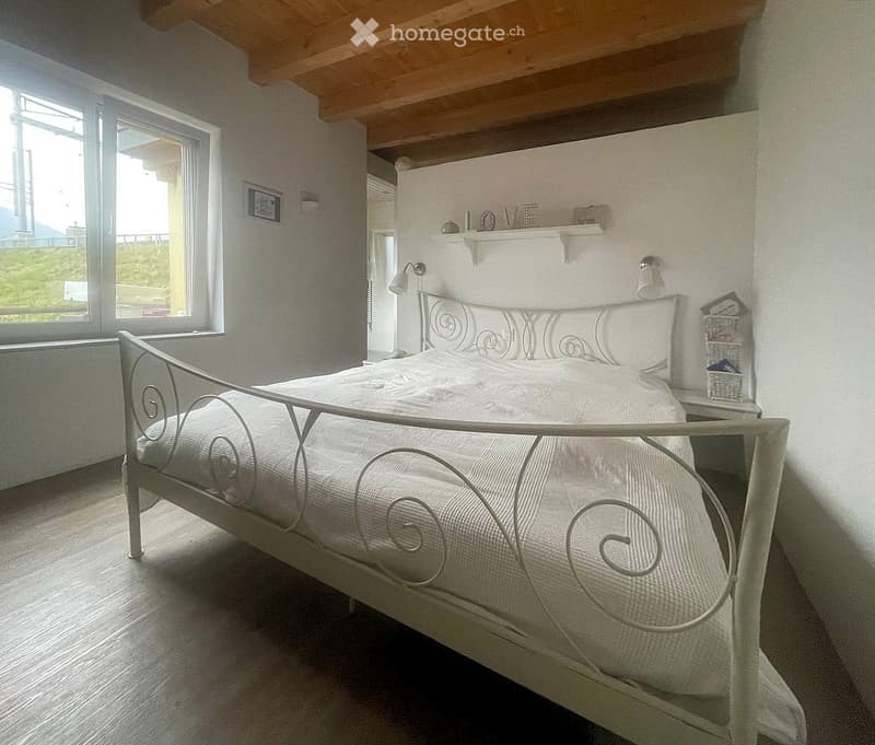 Cozy bedroom with a white metal bed, nightstand, wicker basket, and a shelf with decorative items. Large windows provide natural light.