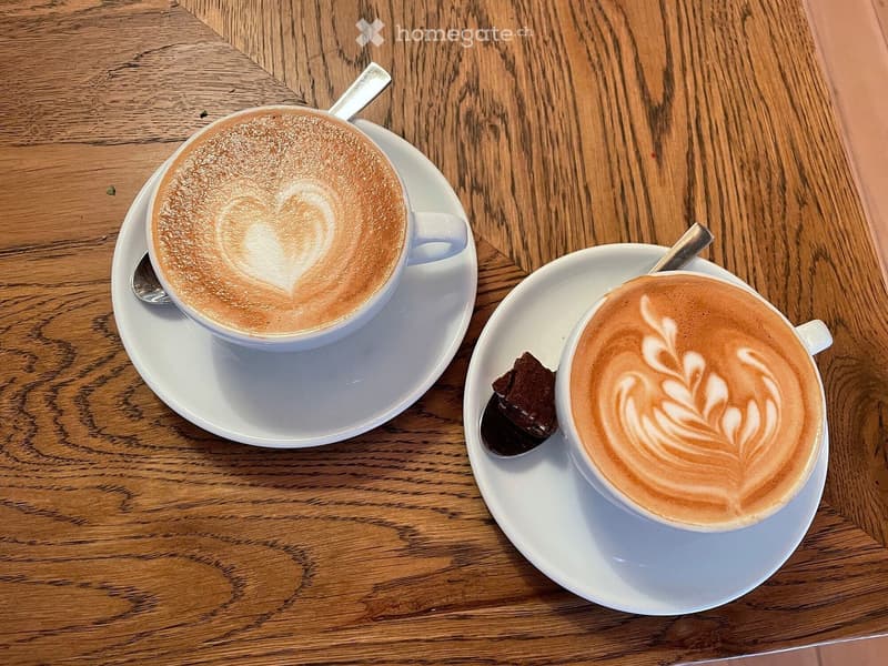 Two cappuccinos with latte art on white saucers, accompanied by a piece of chocolate on a spoon.