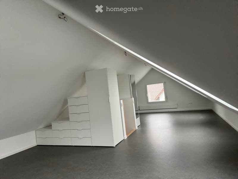 Unfurnished Room. An empty attic room with a slanted ceiling, grey walls, and a single window showing a roof view. The floor is dark grey concrete, and there is a built-in white storage unit with drawers and cabinets on one side.