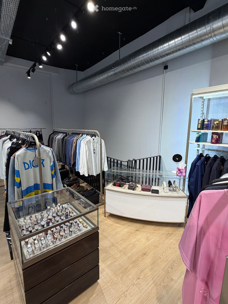 A well-lit retail store with wooden flooring, clothing racks, glass display cases filled with watches and accessories, shelves with perfumes, and a mirror on a stand.