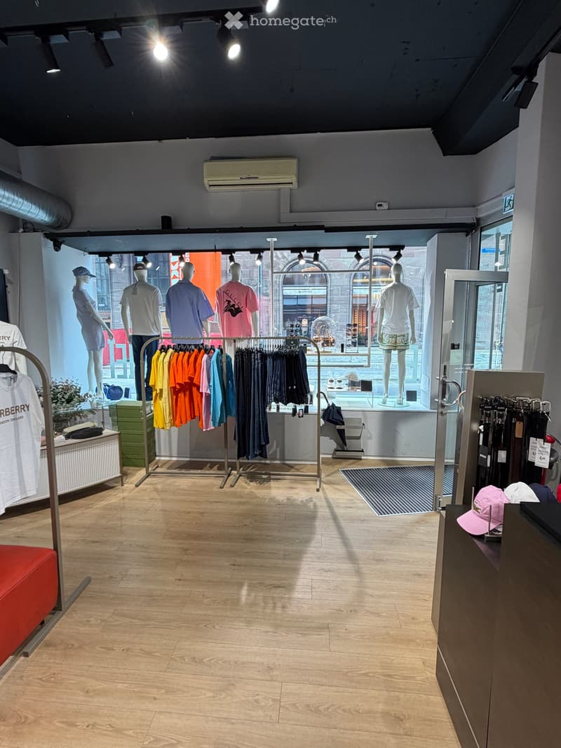 Retail clothing store with wooden flooring, clothing racks, mannequins, and a small seating area.