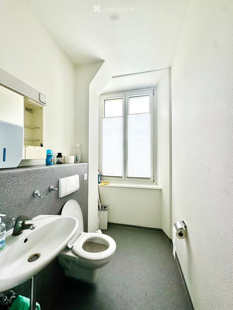 Bathroom with a sink, toilet, mirror, and various toiletries on the counter. The room has a window with blinds and a small trash bin near the window.