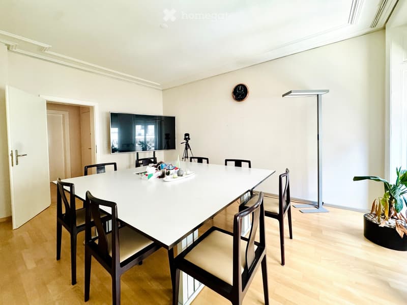 Bright room with a long white table surrounded by chairs, a large screen mounted on the wall, a tripod, a clock, and a potted plant near the window.