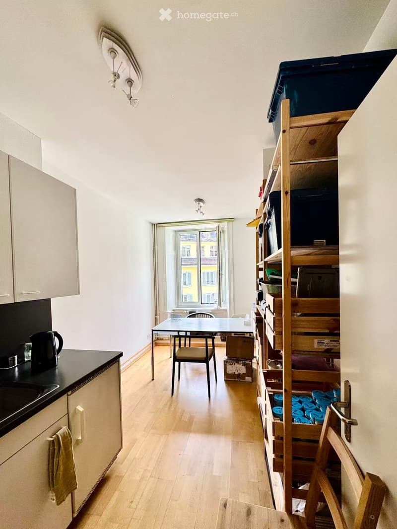 A small, well-lit room with a kitchen area on the left and a dining area in the center. The kitchen features a black countertop, a white cabinet, and a towel hanging from a handle. A small table and chair are positioned near a window that offers a view of