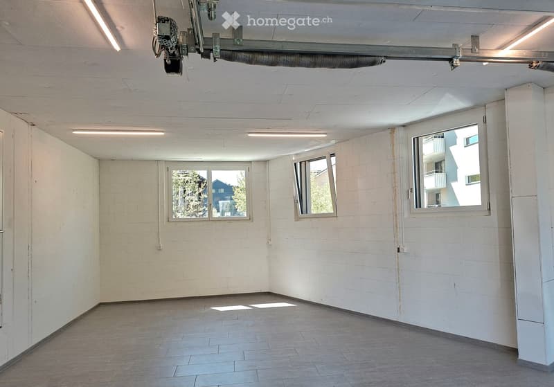 Empty room with white brick walls, tiled floor, and three windows offering views of the outside. The ceiling has fluorescent lighting and a ventilation system.