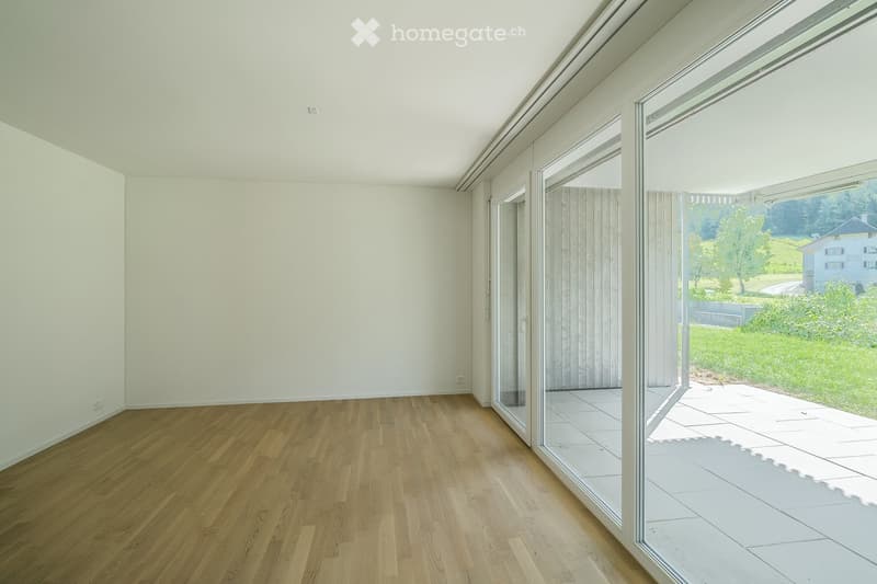 Empty room with wooden flooring and large glass sliding doors leading to a tiled patio area. The walls are white and the ceiling is high.
